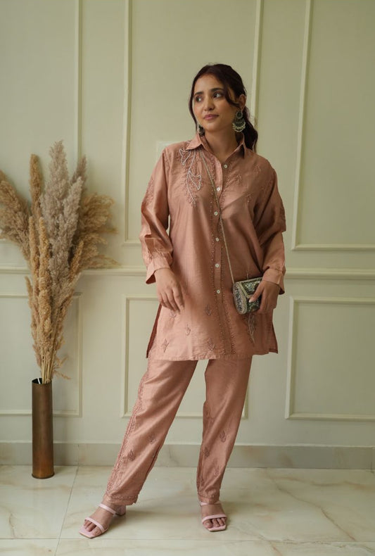 A woman wearing a pink kurta and pants set with intricate embroidery, standing indoors.